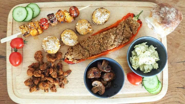 Vegan gef&uuml;llte Paprika und Grillspie&szlig;e sind liebevoll auf einem Holzbrett angerichtet; umgeben von frischen Tomaten und Gurken.
