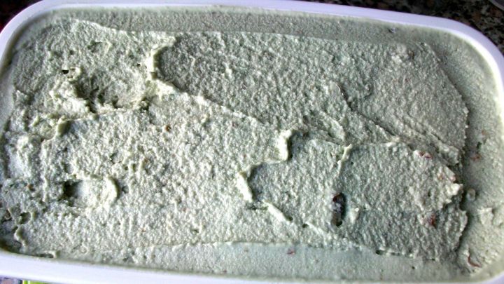 Eine cremige, gr&uuml;ne Avocado-Walnuss-Eiscreme liegt in einem wei&szlig;en Beh&auml;lter auf einem Tisch, umgeben von einer bunten Marmoroberfl&auml;che.