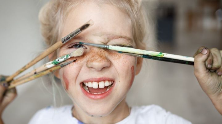 Ein fr&ouml;hliches Kind mit Sommersprossen h&auml;lt Pinsel vor seinem Gesicht und l&auml;chelt, umgeben von einem kreativen Raum.