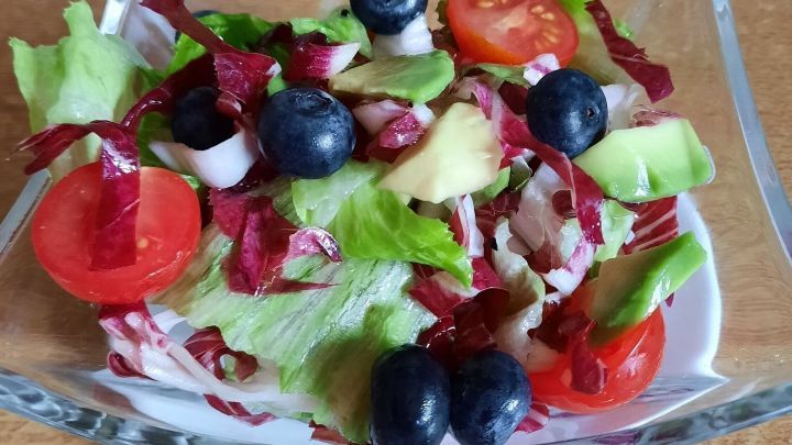 Frischer Salat mit Beeren, Tomaten und Avocado wird in einer klaren Sch&uuml;ssel auf einem Holzuntergrund serviert.