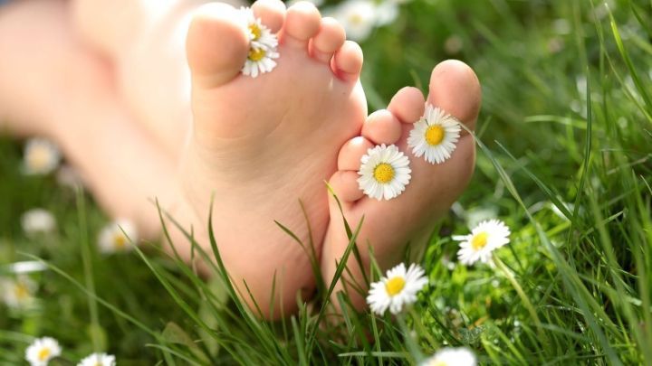 Barfu&szlig;f&uuml;&szlig;e liegen im Gras, geschm&uuml;ckt mit G&auml;nsebl&uuml;mchen, was eine entspannte Atmosph&auml;re der Selbstpflege schafft.