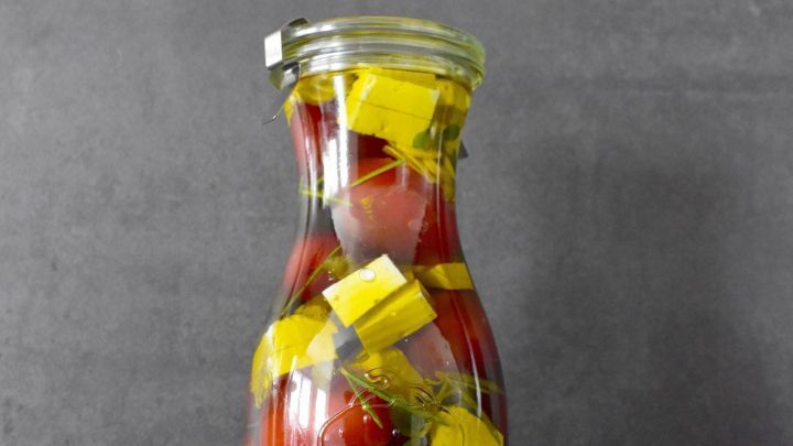 Ein Glas mit eingelegten Cherrytomaten, Feta und Kr&auml;utern steht auf einer dunklen Oberfl&auml;che neben frischem Knoblauch.