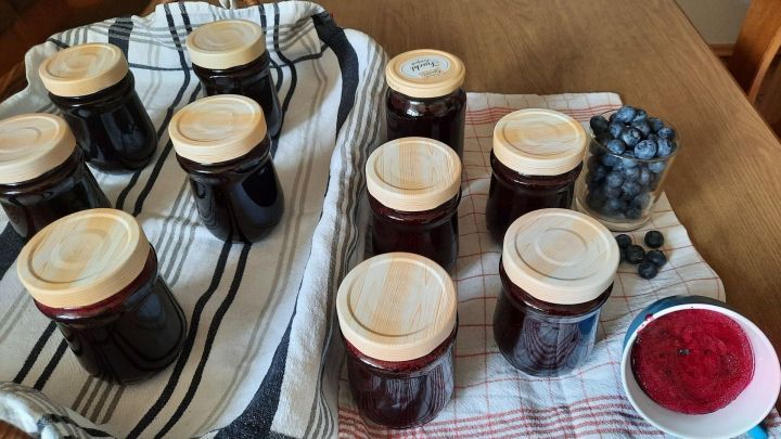 Gl&auml;ser mit Heidelbeermarmelade stehen auf einem Tisch, umgeben von frischen Heidelbeeren und einem Tuch.