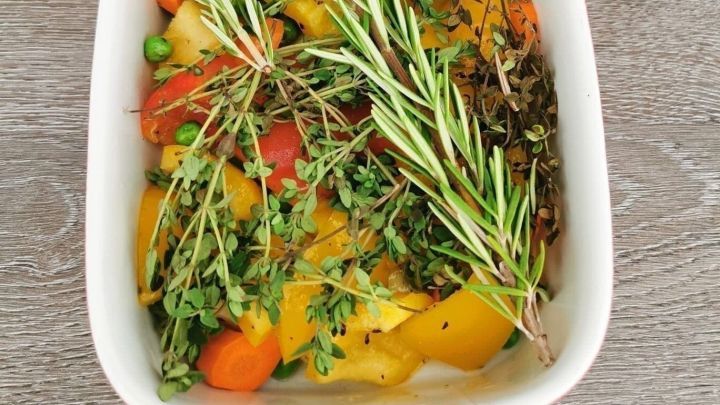 Buntes Ofengem&uuml;se mit frischen Kr&auml;utern ist in einer wei&szlig;en Auflaufform angerichtet, auf einem Holzuntergrund.