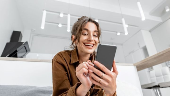 Eine junge Frau l&auml;chelt, w&auml;hrend sie auf ihrem Smartphone liest, in einem hellen, modernen Wohnzimmer.