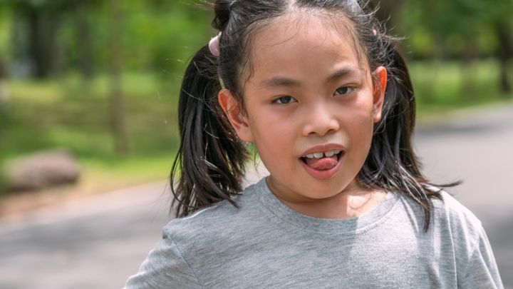 Ein Kind schwitzt und streckt die Zunge heraus, w&auml;hrend es im Park steht. Es tr&auml;gt ein graues T-Shirt und hat Z&ouml;pfe.
