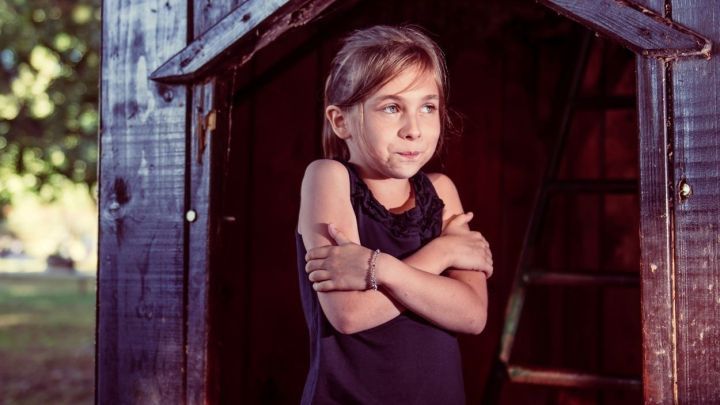 Ein junges M&auml;dchen steht in einem Holzhaus und umklammert sich, w&auml;hrend es leicht fr&ouml;stelt.