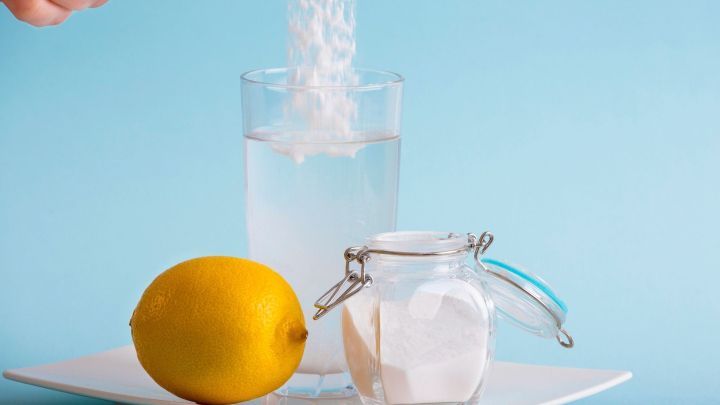 Ein L&ouml;ffel Natron wird in ein Glas Wasser gegeben, w&auml;hrend eine Zitrone und ein offenes Glas Zucker auf einem Teller stehen.