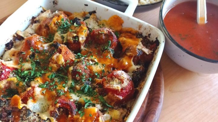 Eine Auflaufform mit gef&uuml;llten Paprika, &uuml;berbacken mit K&auml;se und Kr&auml;utern, steht auf einem Holzbrett, neben Reis und Tomatensuppe.