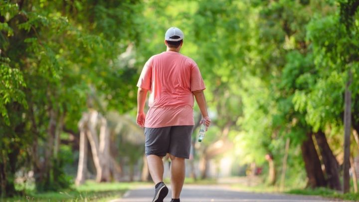 Ein Mann in Sportkleidung geht mit einer Wasserflasche auf einem gr&uuml;nen Weg, umgeben von B&auml;umen und Sonnenlicht.
