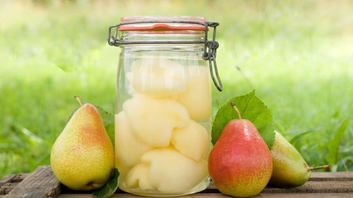 Ein Glas mit eingekochten Birnen steht auf einem Holztisch, umgeben von frischen Birnen und einer gr&uuml;nen Wiese.