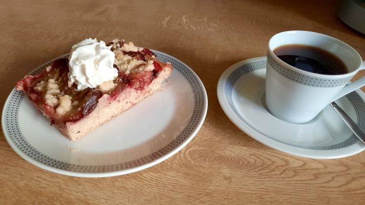 Ein St&uuml;ck Zwetschgenstreuselkuchen mit Sahne liegt auf einem Teller neben einer Tasse Kaffee auf einem Holz Tisch.