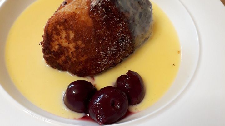 Ein Karth&auml;userkl&ouml;&szlig;e liegt auf einer wei&szlig;en Servierplatte in einer sahnigen So&szlig;e, umgeben von Kirschen und Roter Sauce.