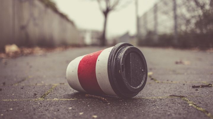 Ein umgefallener Pappbecher liegt auf einem B&uuml;rgersteig, umgeben von tristem, grauem Wetter und kargem Laub.