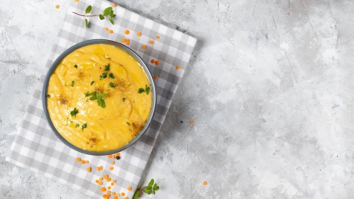 Ein cremiges K&uuml;rbis-Linsen-Curry in einer Sch&uuml;ssel, platziert auf einem karierten Tuch, umgeben von Linsen.