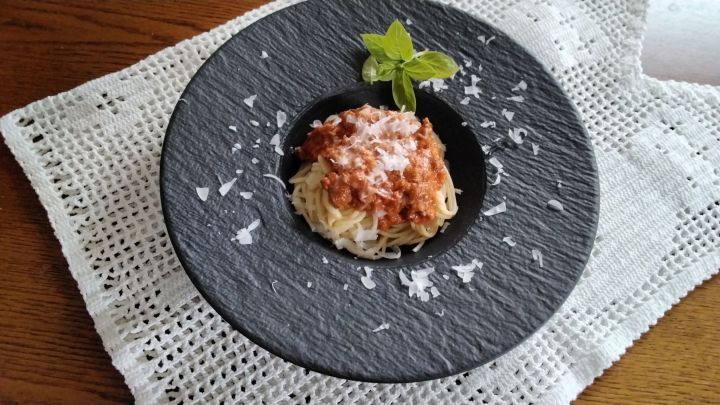 Ein schwarzes Tellergeschirr pr&auml;sentiert Spaghetti mit Tomaten-Hackfleisch-So&szlig;e, garniert mit Basilikum und K&auml;se.