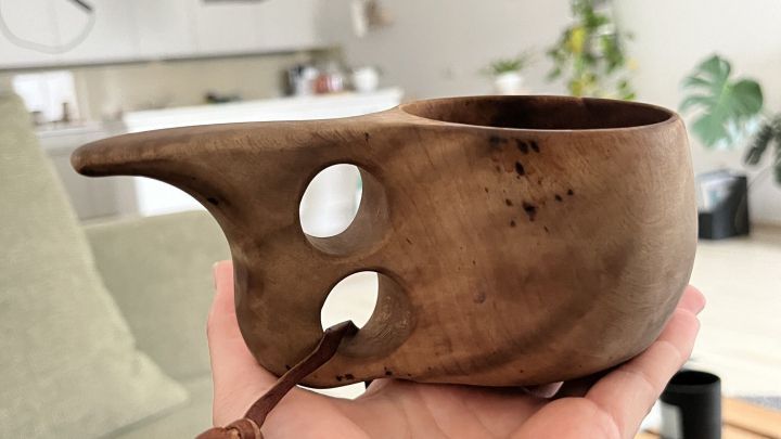 Eine handgefertigte Kuksa wird in einer Hand gehalten, umgeben von einer modernen Wohnk&uuml;che mit Pflanzen und Dekoration.
