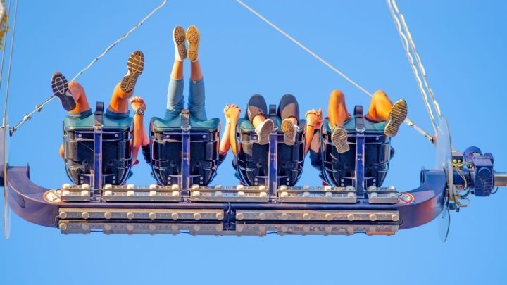 F&uuml;nf Personen sitzen kopf&uuml;ber in einem Karussell, w&auml;hrend sie an den Sicherheitsvorrichtungen festhalten, unter blauem Himmel.