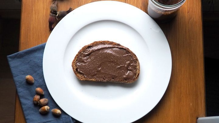 Ein St&uuml;ck Brot liegt auf einem Teller, bestrichen mit Nougat, umgeben von Haseln&uuml;ssen und einem Glas Inhalt.
