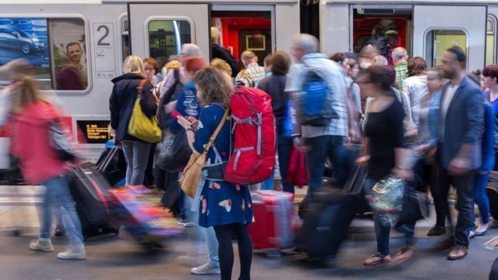 Eine Frau mit rotem Rucksack steht mit ihrem Handy in der Menge am Bahnhof, w&auml;hrend Reisende in den Zug einsteigen.