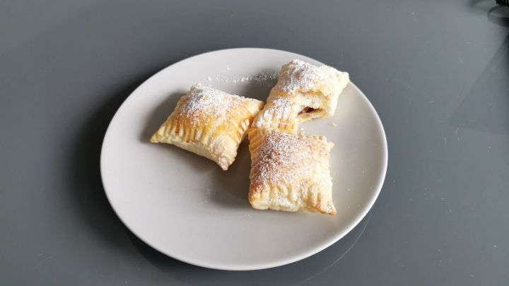 Drei Bl&auml;tterteigtaschen liegen auf einem wei&szlig;en Teller, bestreut mit Puderzucker, bereit zum Genie&szlig;en in einem hellen Raum.