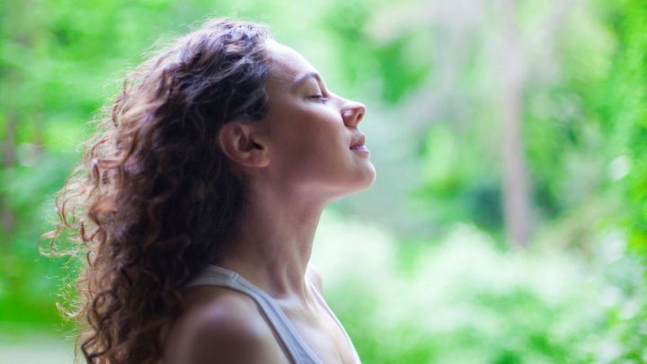 Eine Frau atmet tief ein, w&auml;hrend sie in einer gr&uuml;nen, ruhigen Umgebung steht, um Stress und Angst abzubauen.