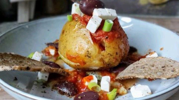 Eine gef&uuml;llte Ofenkartoffel steht mittig auf einem Teller, dekoriert mit Tomatensauce, Feta, Oliven und Brot.