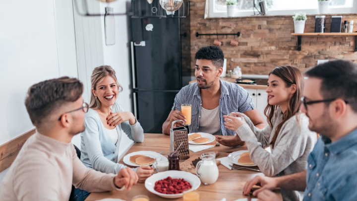 Sechs junge Erwachsene genie&szlig;en ein Fr&uuml;hst&uuml;ck am Tisch. Lachen und Gespr&auml;che f&ouml;rdern ein harmonisches Zusammenleben.