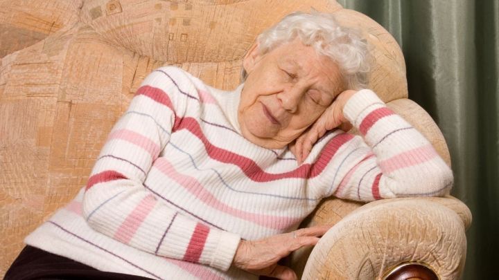 Eine &auml;ltere Frau schl&auml;ft auf einem Sofa, w&auml;hrend sie gem&uuml;tlich in einem gestreiften Pullover sitzt, umgeben von einem ruhigen Raum.