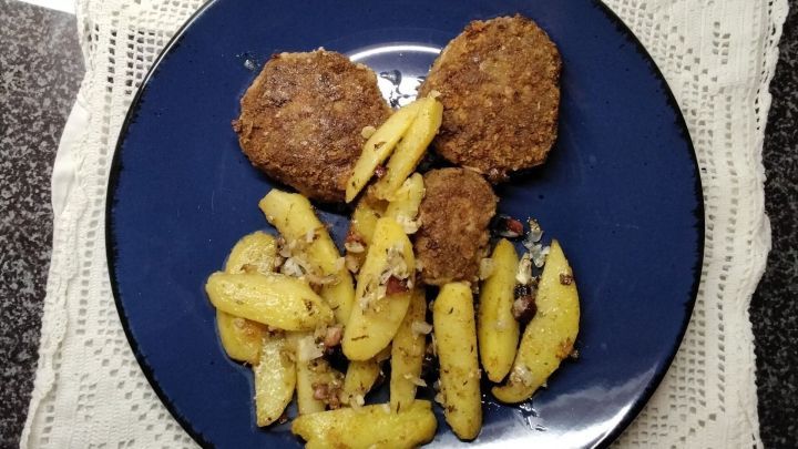 Frikadellen liegen neben goldbraun gebratenen Kartoffeln auf einem blauen Teller, umgeben von einer wei&szlig;en Tischdecke.