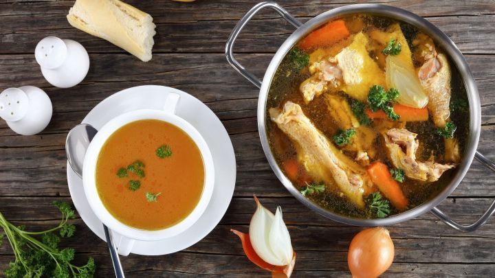 Eine dampfende H&uuml;hnerbr&uuml;he kocht in einem Topf, w&auml;hrend eine Schale mit klarer Br&uuml;he neben frischem Brot auf einem Holztisch steht.