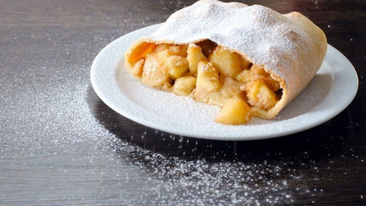Auf einem wei&szlig;en Teller liegt ein Apfelstrudel mit sichtbar herausschauenden &Auml;pfeln, garniert mit Puderzucker auf einem dunklen Tisch.