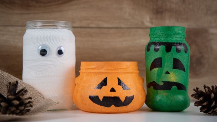 Bunte Halloween-Windlichter aus Altglas zeigen eine gruselige Atmosph&auml;re auf einem Holzuntergrund, umgeben von Naturmaterialien.