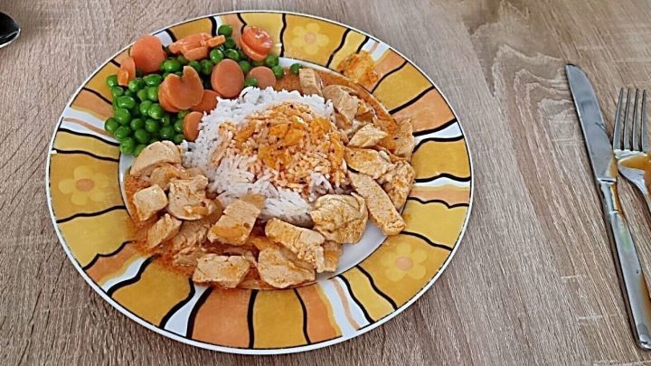 Ein bunter Teller mit H&auml;hnchengeschnetzeltem und Reis, begleitet von Erbsen und Karotten, auf einem holzfarbigen Tisch.