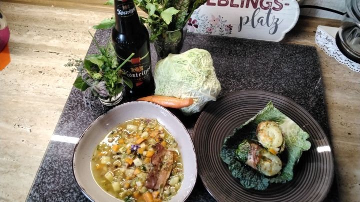 Gef&uuml;llte Wirsingrouladen liegen auf einem Teller neben einer deftigen Gem&uuml;sesuppe und frischen Zutaten auf einem Tisch.