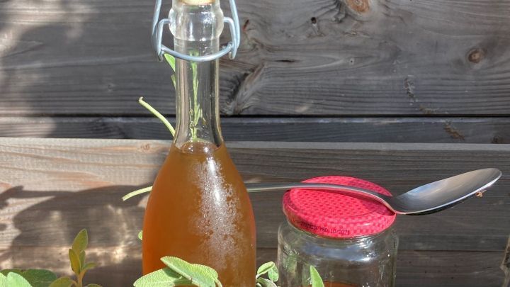Eine Flasche mit selbstgemachtem Hustensaft steht zwischen gr&uuml;nen Kr&auml;utern auf einem Holztisch, eine L&ouml;ffel ruht daneben.