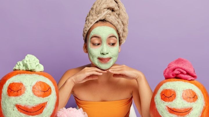 Eine Frau mit einer Gesichtsmaske l&auml;chelt charmant zwischen zwei dekorierten K&uuml;rbissen, die Fruchtbarkeit im Herbst symbolisieren.