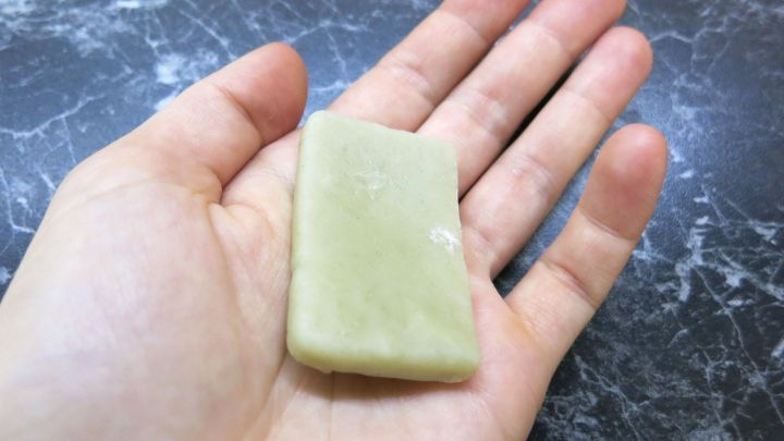 Eine Hand h&auml;lt ein St&uuml;ck Olivenseife, das leicht gl&auml;nzt, auf einer dunklen, marmorierten Oberfl&auml;che.