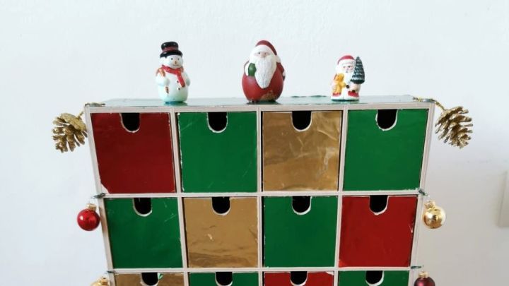 Ein Adventskalender mit Holzschubladen steht dekoriert auf einem Tisch, geschm&uuml;ckt mit Weihnachtsfiguren und Kugeln.