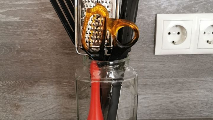 Eine Glasbeh&auml;lter steht auf einer Arbeitsfl&auml;che und h&auml;lt K&uuml;chenutensilien wie einen R&uuml;hrbesen und eine Reibe.