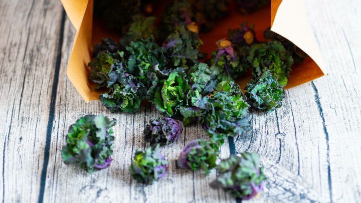Kale-Bl&uuml;ten fallen aus einer braunen T&uuml;te auf einen rustikalen Holztisch und zeigen ihre gr&uuml;ne und violette F&auml;rbung.