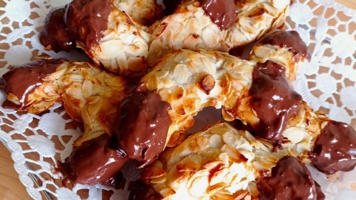 Auf einem wei&szlig;en, filigranen Teller liegen frisch gebackene Mandelh&ouml;rnchen, zur H&auml;lfte in Schokolade getaucht.