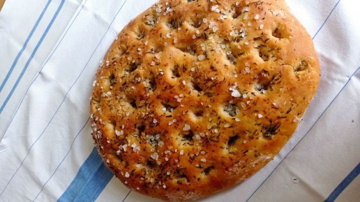 Eine goldbraun gebackene Focaccia liegt auf einem wei&szlig;en Tisch mit blauen Streifen, bestreut mit grobem Salz und Kr&auml;utern.