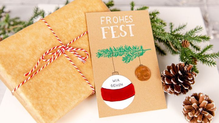 Ein festlich dekoriertes Rubbellos auf einem Geschenk, umgeben von Tannenzweigen und Tannenzapfen, bringt Weihnachtsstimmung.