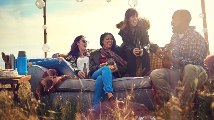 Eine Gruppe von Freunden sitzt entspannt auf einer Couch im Freien, lacht, h&auml;lt Getr&auml;nke und genie&szlig;t eine herbstliche Atmosph&auml;re.