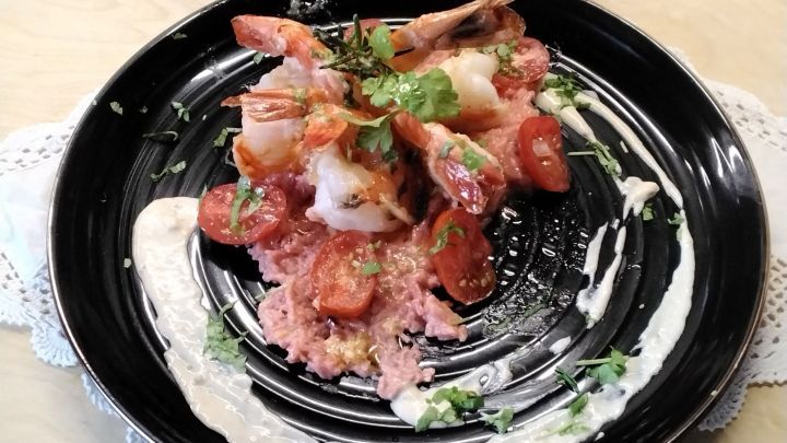 Schwarz-gelbe Garnelen liegen auf rosa Risotto, garniert mit Tomaten und Kr&auml;utern, umgeben von cremiger So&szlig;e.