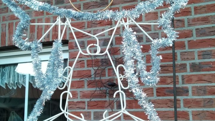 Ein silberner Engel aus Kleiderb&uuml;geln h&auml;ngt an einer Wand aus Ziegelsteinen und ist mit Lametta dekoriert.