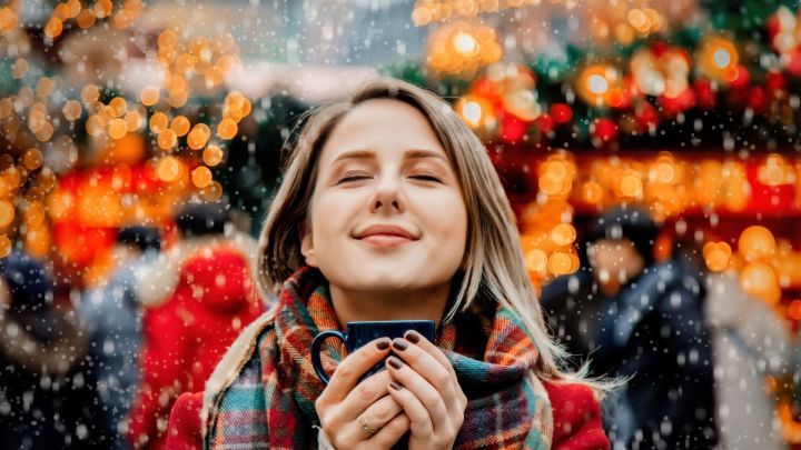 Eine l&auml;chelnde Frau genie&szlig;t einen warmen Becher inmitten eines festlich beleuchteten Weihnachtsmarkts im Schnee.