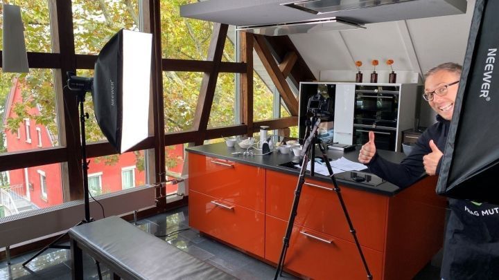 Ein Mann zeigt Daumen hoch, w&auml;hrend er in einer modernen, orangefarbenen K&uuml;che mit Kamera und Lichtausr&uuml;stung posiert.