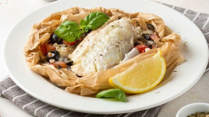 Gegrillter Seelachs liegt in einem Gem&uuml;se-P&auml;ckchen, umgeben von frischen Zitronenscheiben und Kr&auml;utern auf einem Teller.